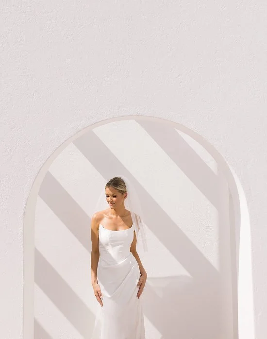 A woman in a white dress standing in front of a arched opening with shadows on a white wall.