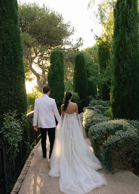 Bride and groom walking hand in hand on a garden pathway surrounded by tall trees and shrubs during sunset.