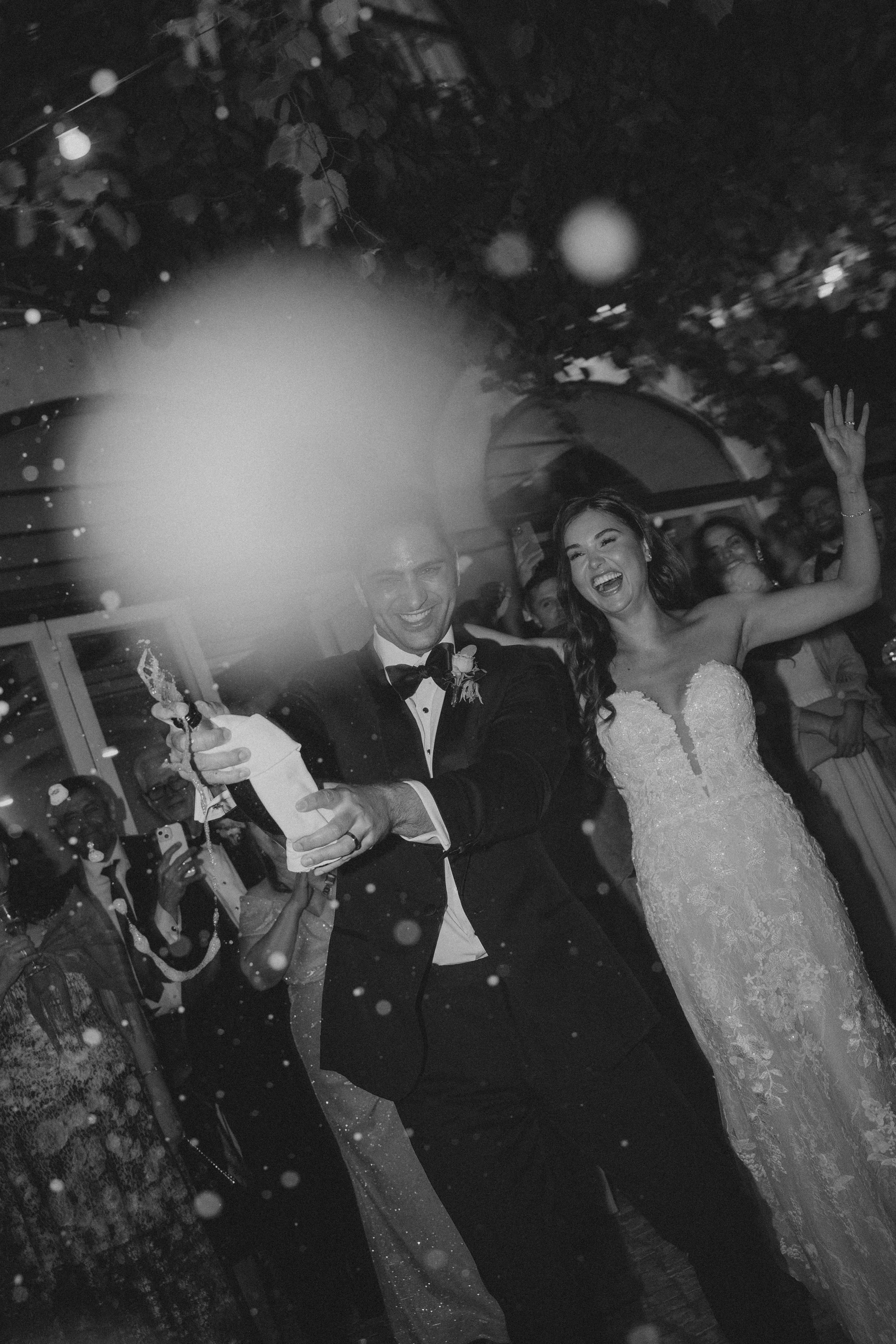 A bride and groom dancing at their wedding reception, smiling and celebrating, in a black and white photo with guests in the background.