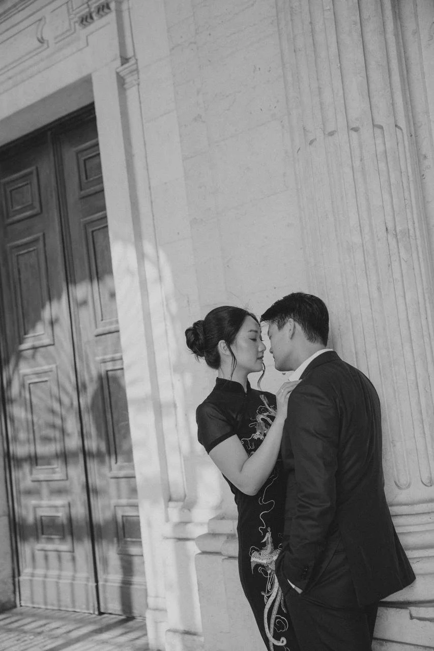 A black and white photo of a man and woman standing close, about to kiss, in front of a building with large columns and a wooden door, with shadows of branches on the wall.