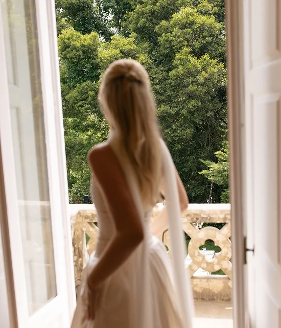 A woman in a white dress standing at an open balcony door, looking outside at green trees.