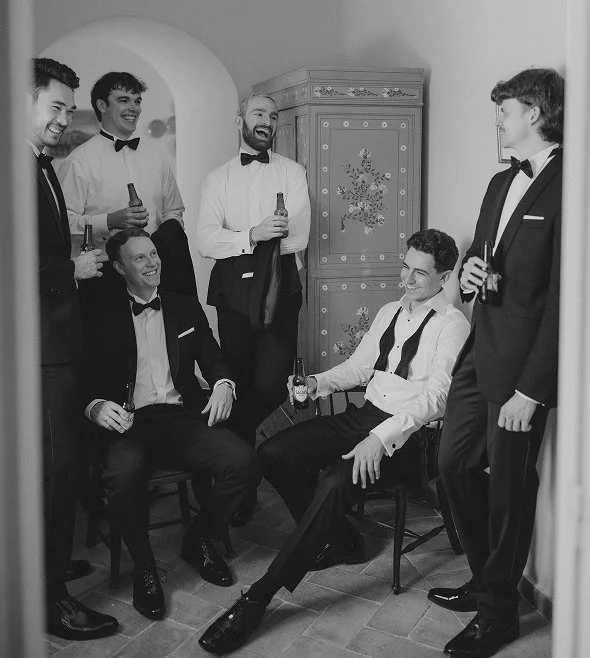 Group of six men in tuxedos and bow ties socializing indoors; one seated with unbuttoned shirt and loosened tie, smiling, two seated and four standing, all with drinks, in a room with a decorative cabinet.