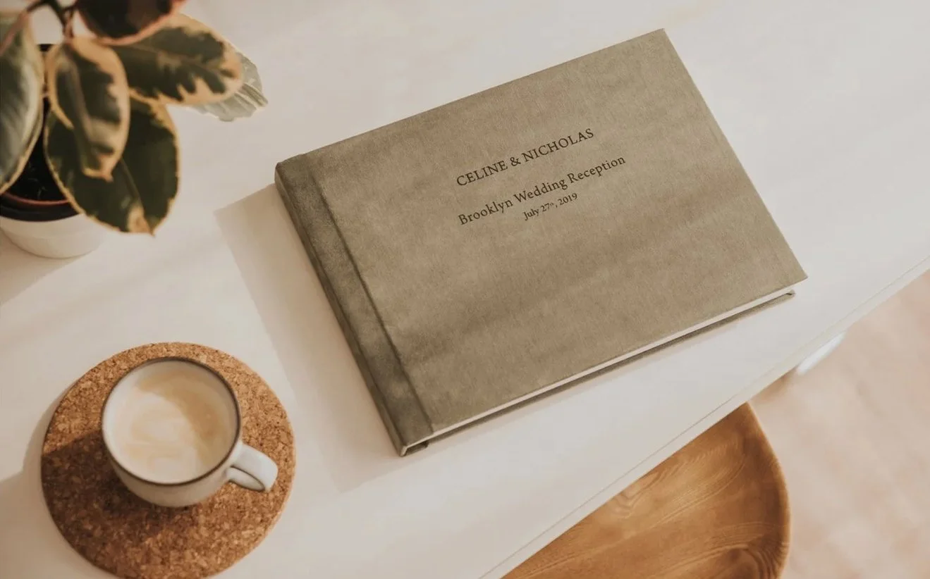 Wedding invitation album titled 'Celina & Nicholas Brooklyn Wedding Reception July 27, 2019' on a white table with a potted plant and a cup of coffee on a cork coaster nearby.
