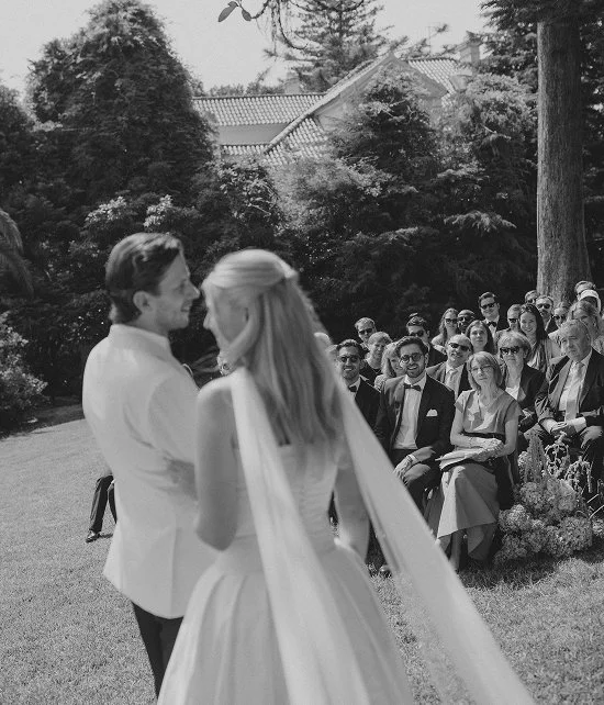 A bride and groom holding hands during their outdoor wedding ceremony with guests seated on the grass behind them.