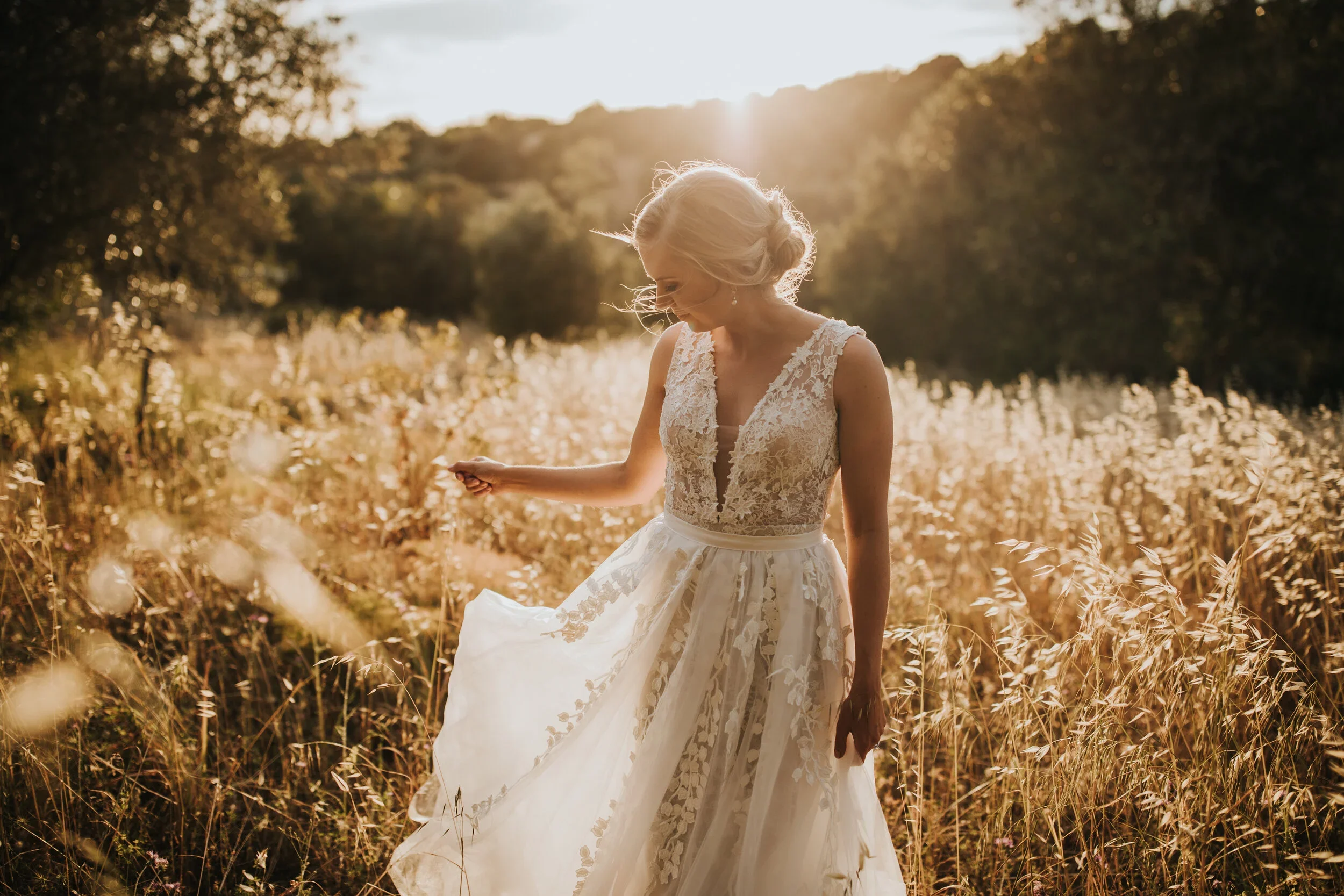A woman in a white lace wedding dress standing in a field of tall grass during sunset with sunlight behind her.