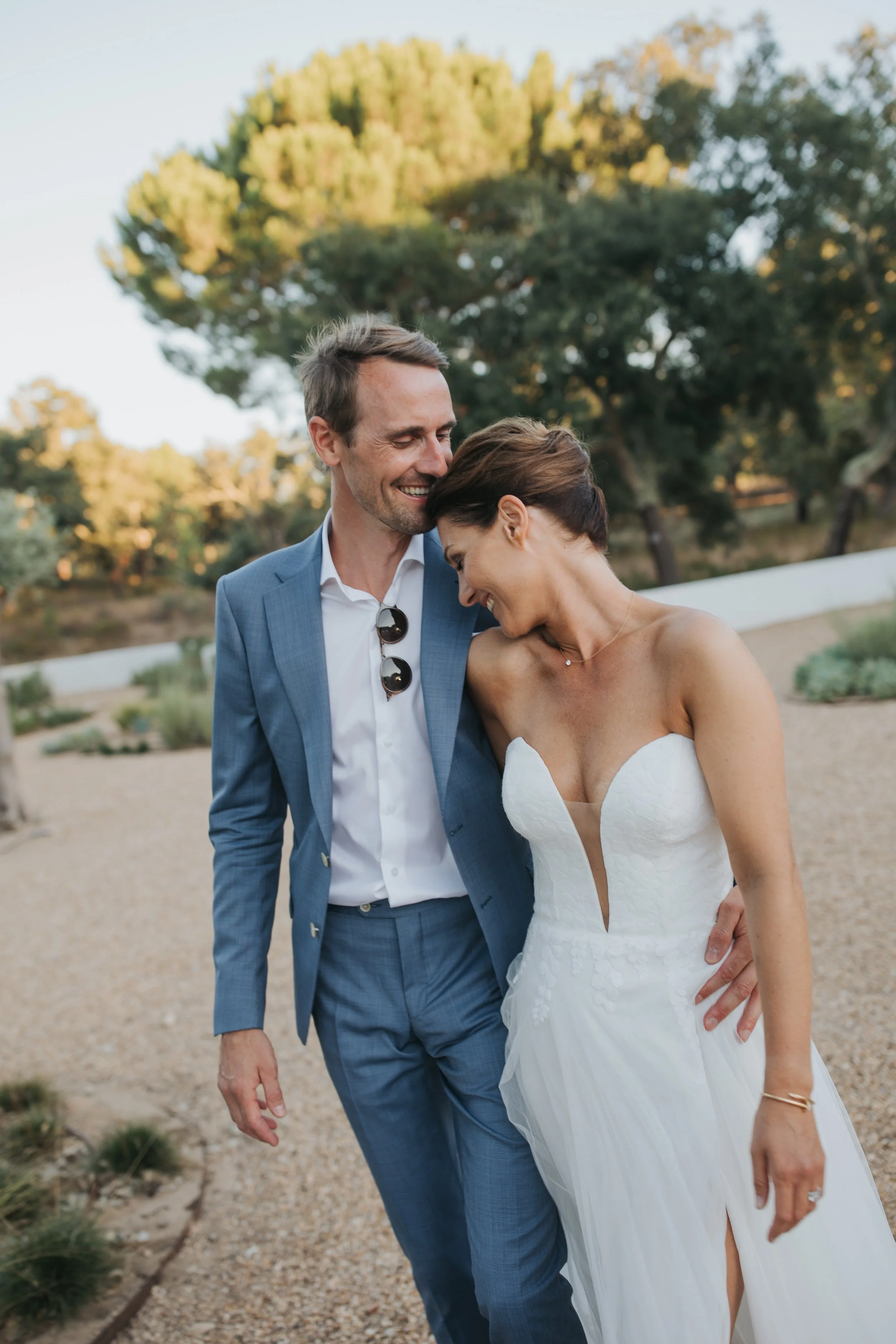 A happy couple in wedding attire sharing a moment outdoors during sunset, with trees and a gravel path in the background.