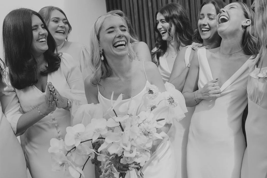 Group of women in white dresses laughing and smiling, with one woman holding a bouquet of flowers, at a wedding celebration.