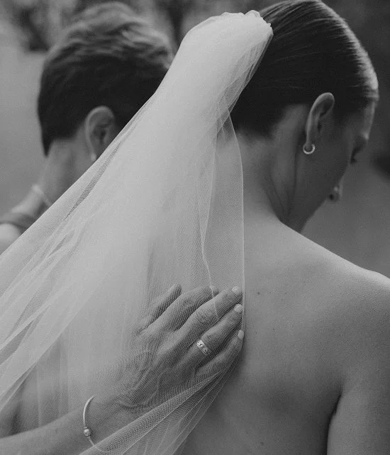 A close-up black and white photo of a bride with a veil and an earring, embracing another person, with a hand placed on her shoulder.