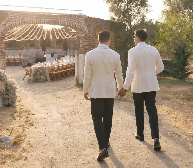 Two men wearing white tuxedos holding hands and walking on a dirt path towards an outdoor wedding reception area with a long table and chairs, decorated with string lights, at sunset.