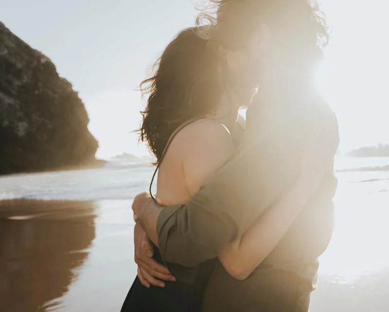 A couple embracing on a beach at sunset with the sun shining brightly behind them.