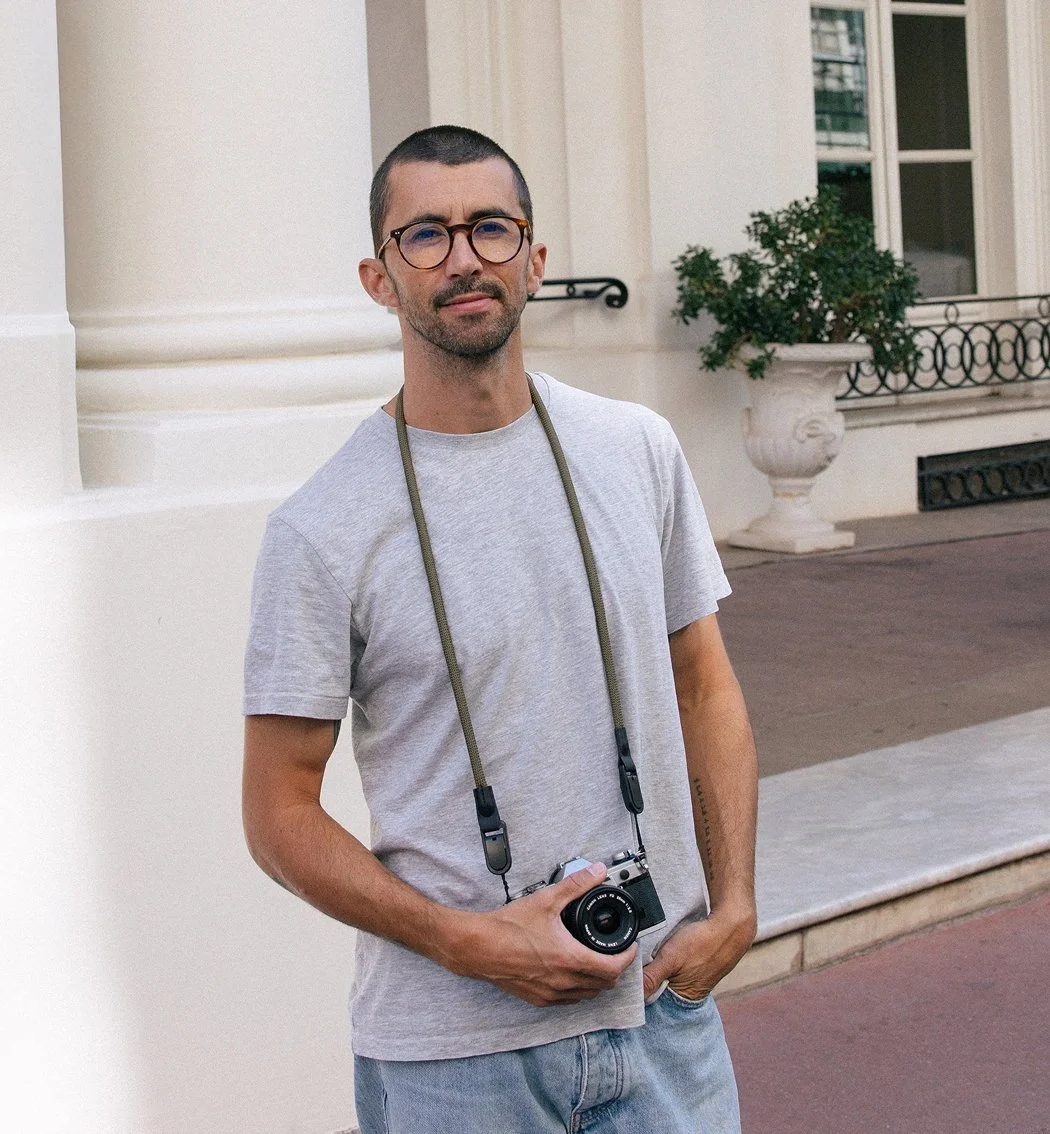 Hugo Coelho holding a camera, standing outdoors in front of a building with columns and a decorative balcony.