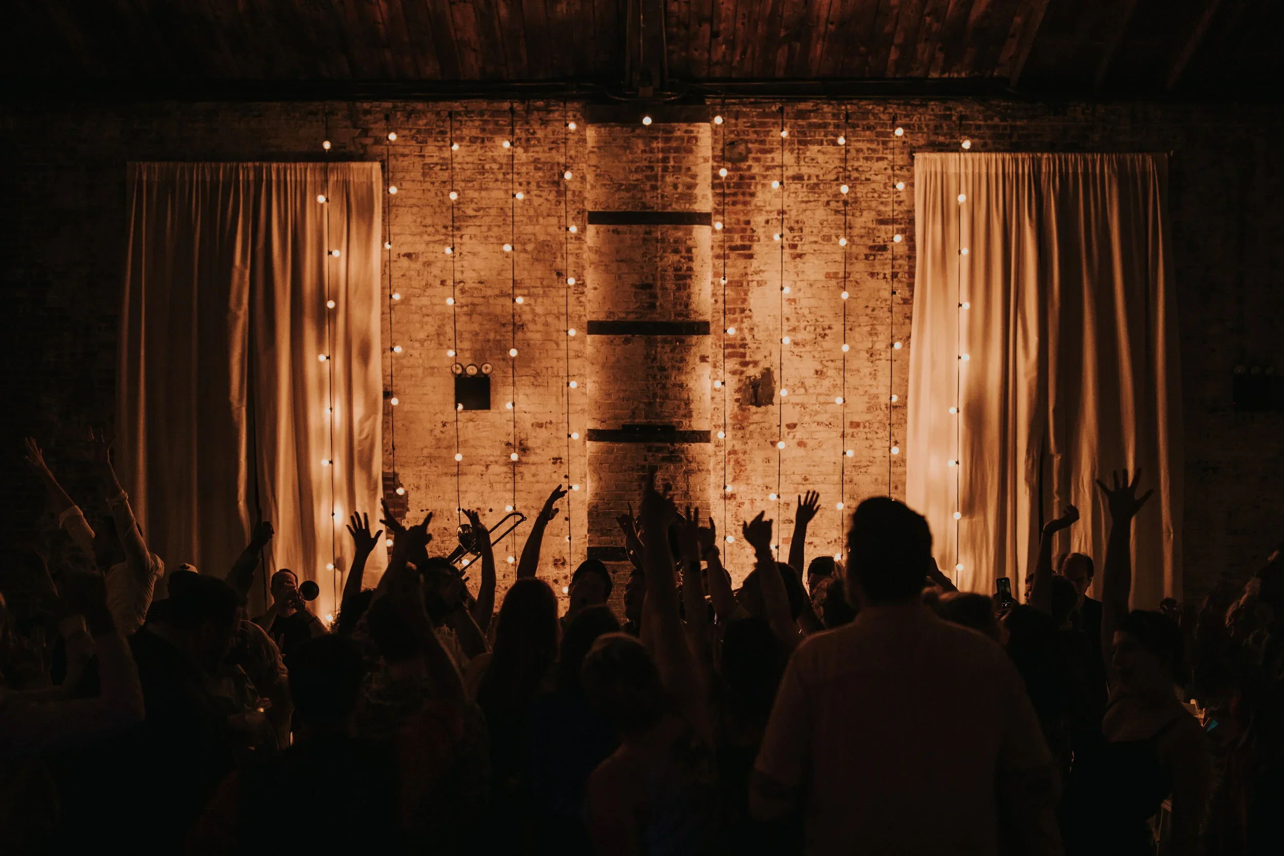 Crowd attending a concert or performance in an indoor venue with exposed brick walls and string lights.