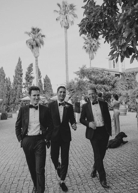 Three men in tuxedos walking outdoors at a formal event, smiling, with palm trees and other guests in the background.
