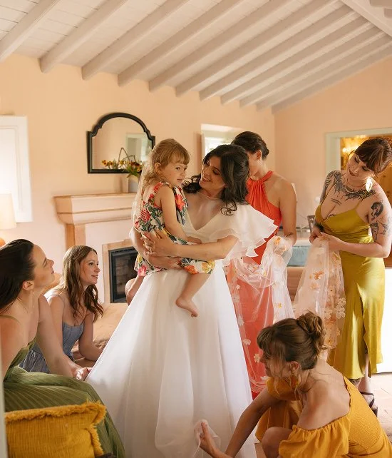 A bride in a white dress holding a young girl in colorful clothing, surrounded by women in bright dresses, smiling and chatting indoors.