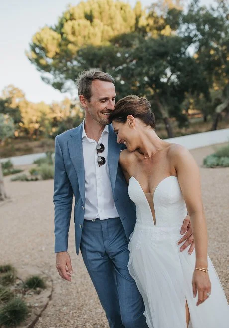 A happy couple, a man in a blue suit and a woman in a white wedding dress, standing outdoors on a gravel path surrounded by trees. They are smiling and leaning close together, sharing an intimate moment.