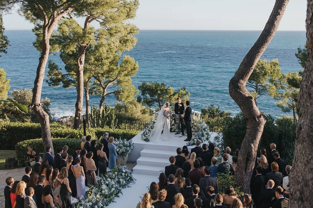 A wedding ceremony taking place outdoors near the ocean, with a bride and groom standing on a white platform, surrounded by guests and decorated with flowers, trees, and ocean scenery in the background.