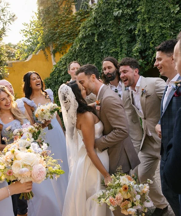 A bride and groom share a kiss during their wedding surrounded by friends and bridesmaids holding bouquets.