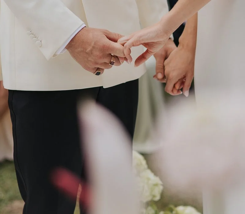 Close-up of a couple holding hands during a wedding ceremony, with the groom wearing a white tuxedo jacket and the bride's hand gently gripping his.