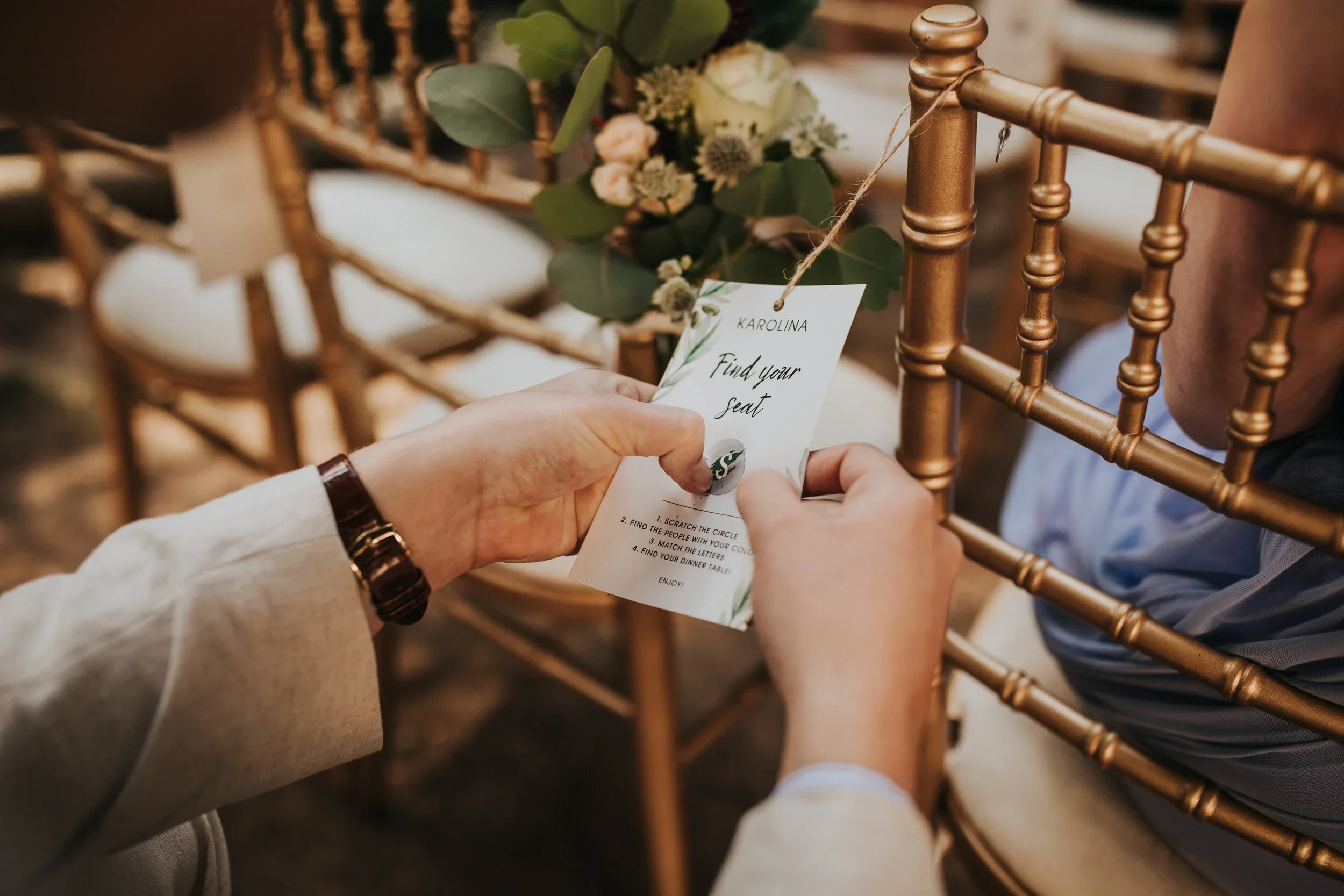 A person holding a 'Find Your Seat' card at a wedding or event, with gold chairs and floral decorations in the background.