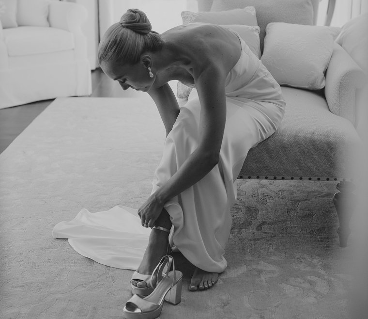 A woman in a strapless dress sitting on a sofa, adjusting her high-heeled shoe on her foot.