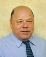 A middle-aged man with a receding hairline, wearing a light blue dress shirt and a dark blue tie, standing against a neutral background.
