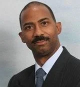 Portrait of a man with a bald head and mustache, wearing a dark suit and tie, against a plain background.