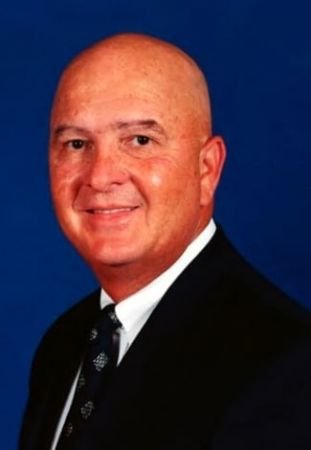 A portrait of a bald man wearing a black suit, white shirt, and patterned tie, smiling against a blue background.