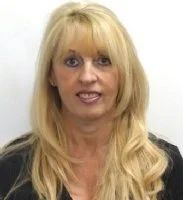 A woman with long blonde hair and bangs, wearing a black top, looking at the camera.