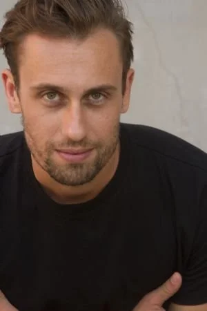 Close-up of a man with short brown hair, a beard, and blue eyes, wearing a black shirt, looking directly at the camera.