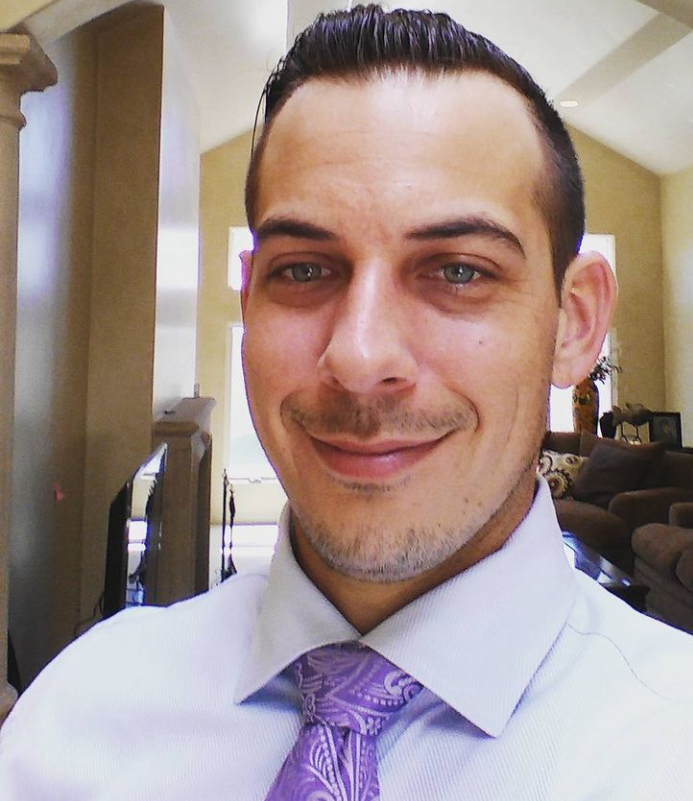 A man with slicked-back dark hair and a trimmed goatee wearing a light-colored shirt and purple tie, smiling indoors with a living room in the background.