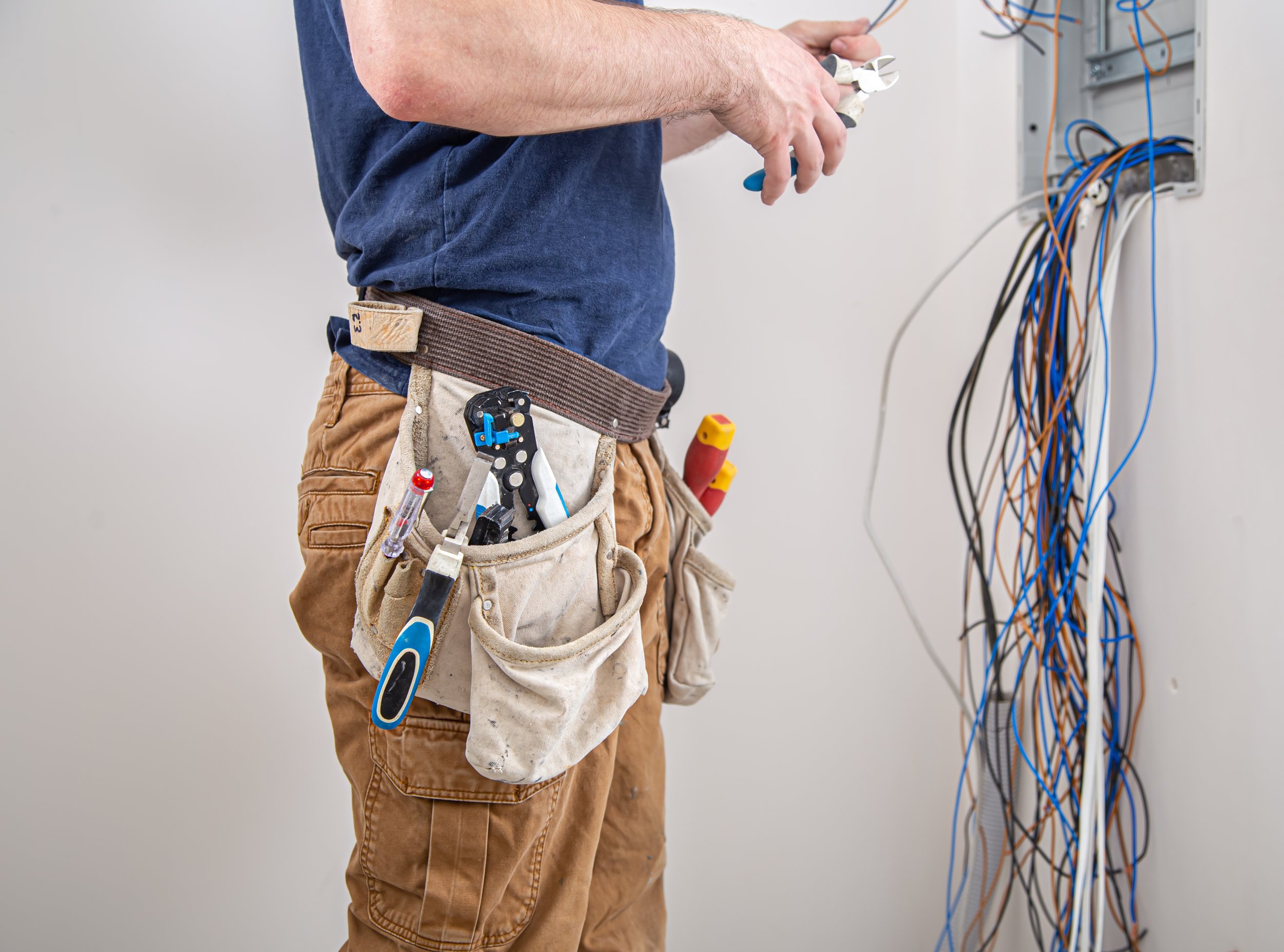 Un homme en jeans et t-shirt travaille avec des outils de câblage électrique devant un panneau électrique avec câbles colorés.