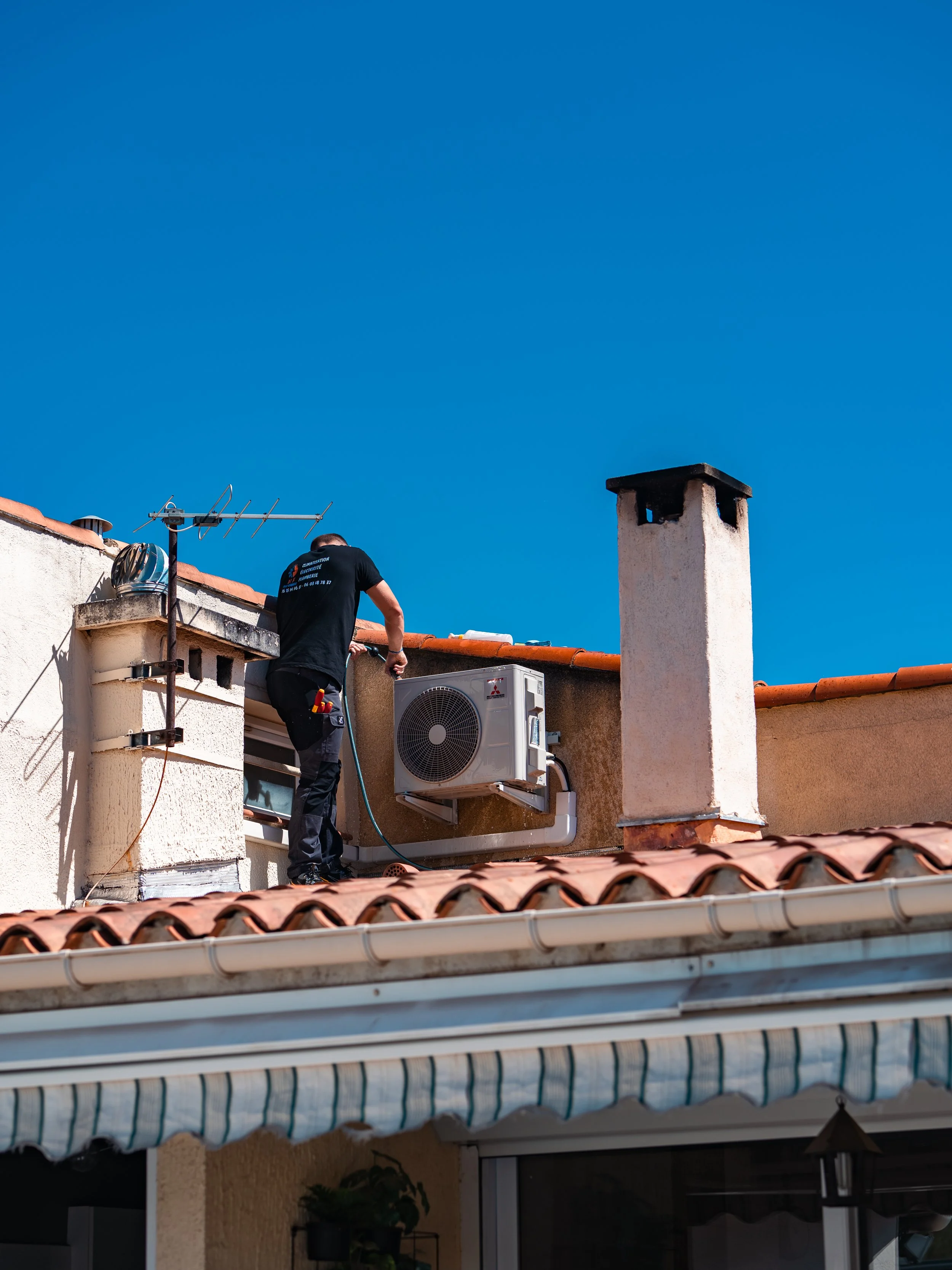 Un homme effectue des travaux de réparation ou d'entretien sur un toit d'immeuble, avec un climatiseur, une antenne et une cheminée visibles, sous un ciel bleu clair.