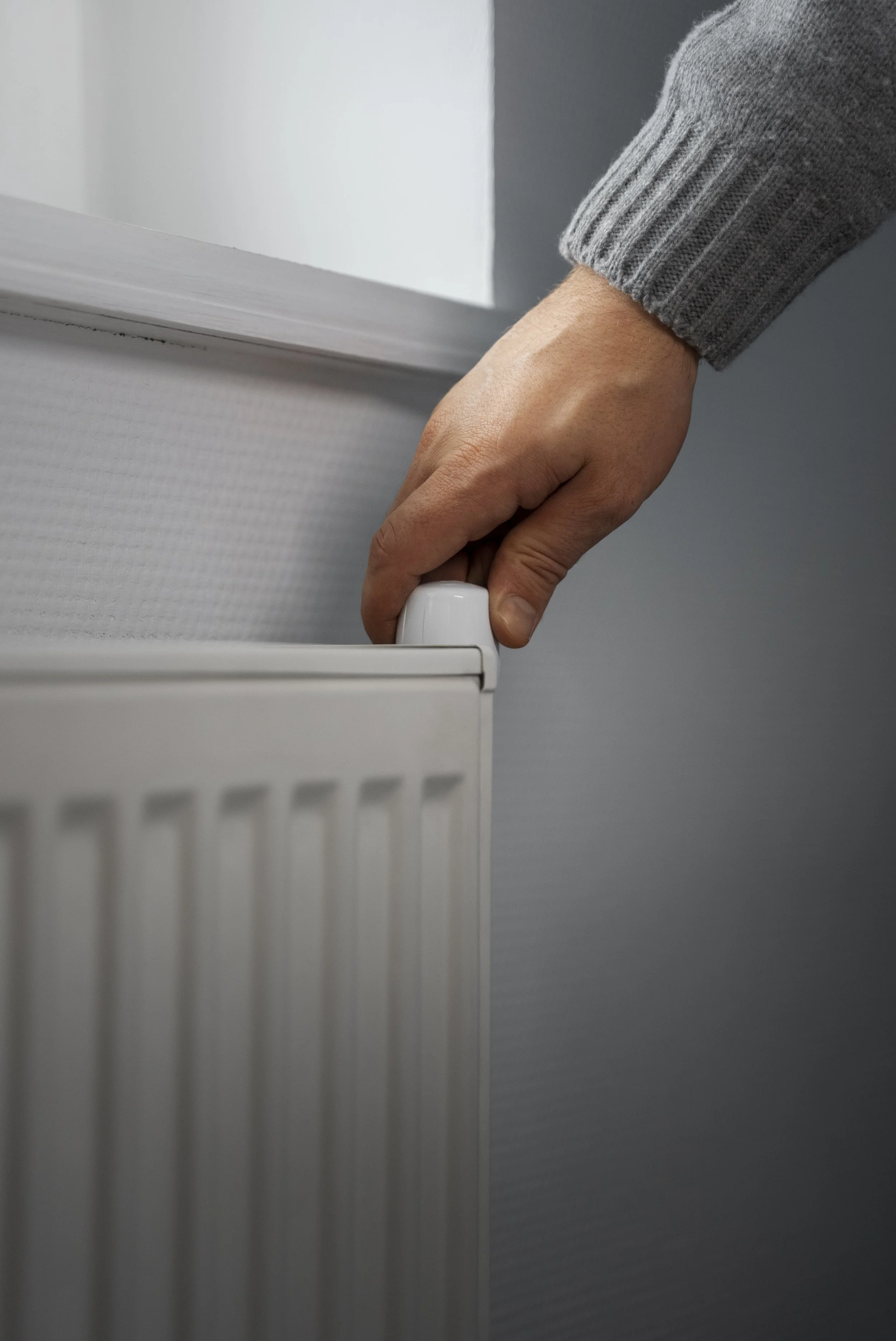 Une main ajustant la valve d'un radiateur électrique blanc, vêtu d'un pull gris à manches longues, dans une pièce à murs gris et blanc.