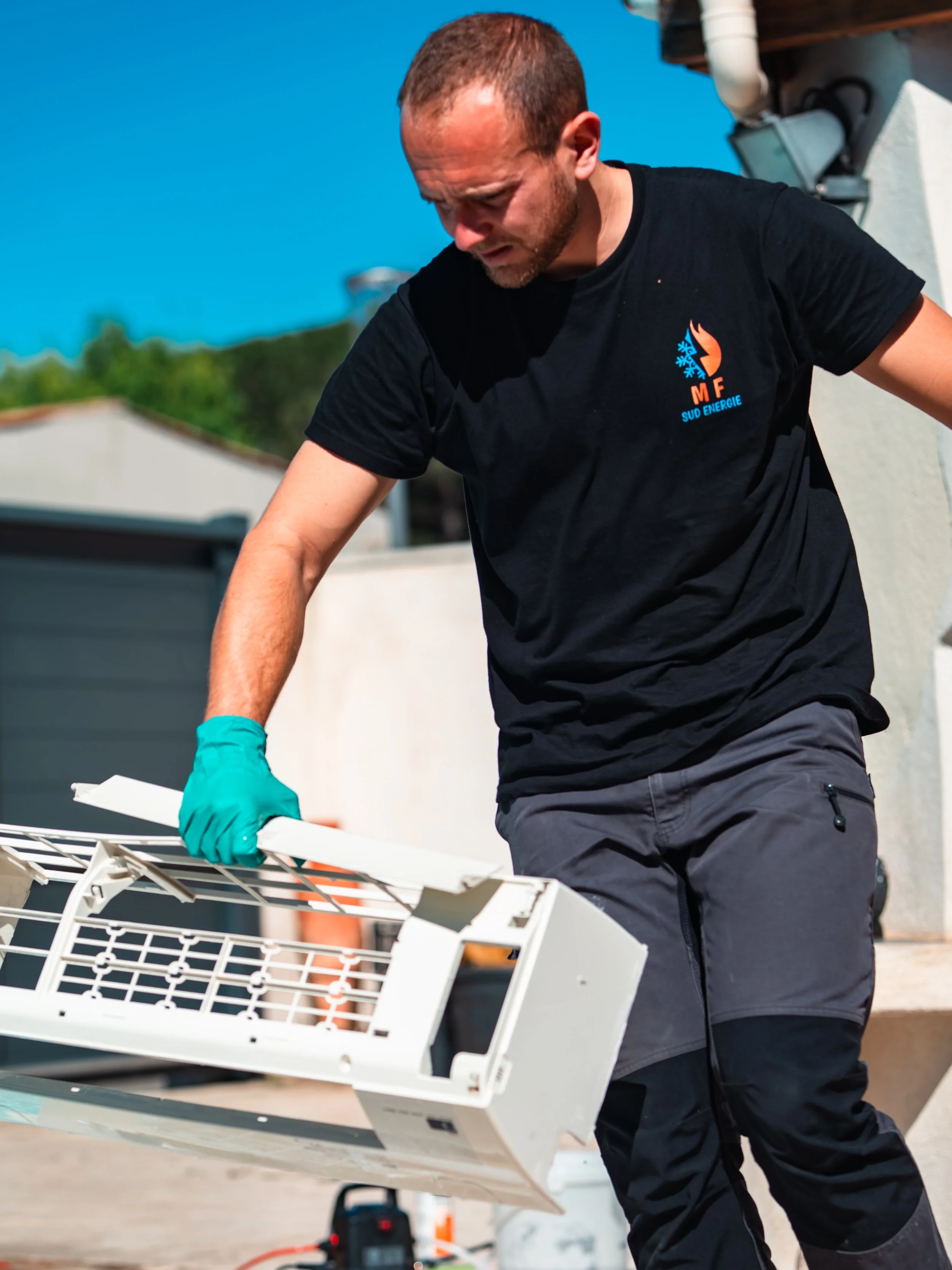 Un homme portant un t-shirt noir avec un logo, manipulant une climatisation lors d'une activité extérieure.