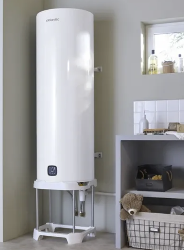 Un chauffe-eau vertical blanc placé dans une pièce avec un meuble à côté contenant des articles de bain et une fenêtre avec des bouteilles et un pot sur le rebord.