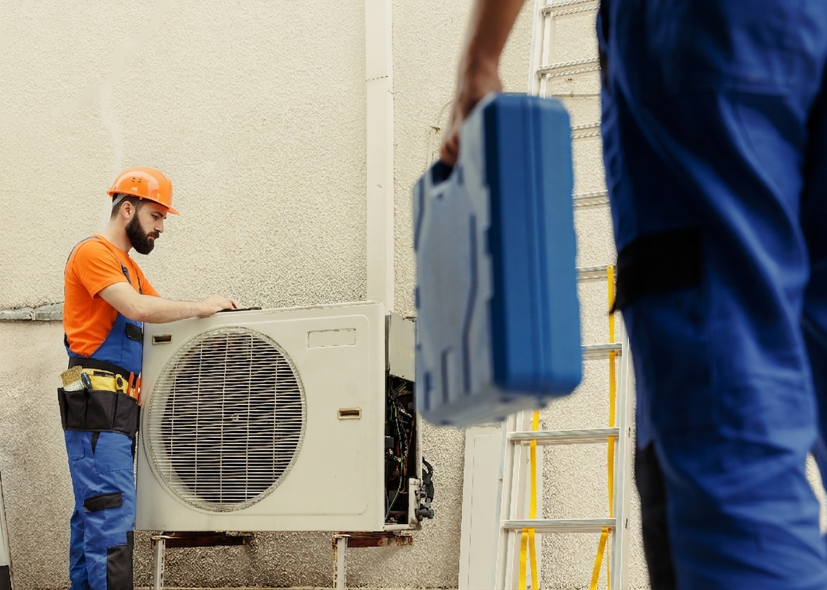 Un technicien travaille à l'entretien d'une unité de climatisation extérieure. Il porte un casque orange et un uniforme bleu et orange, tenant un outil, avec un partenaire en bleu tenant une boîte à outils.