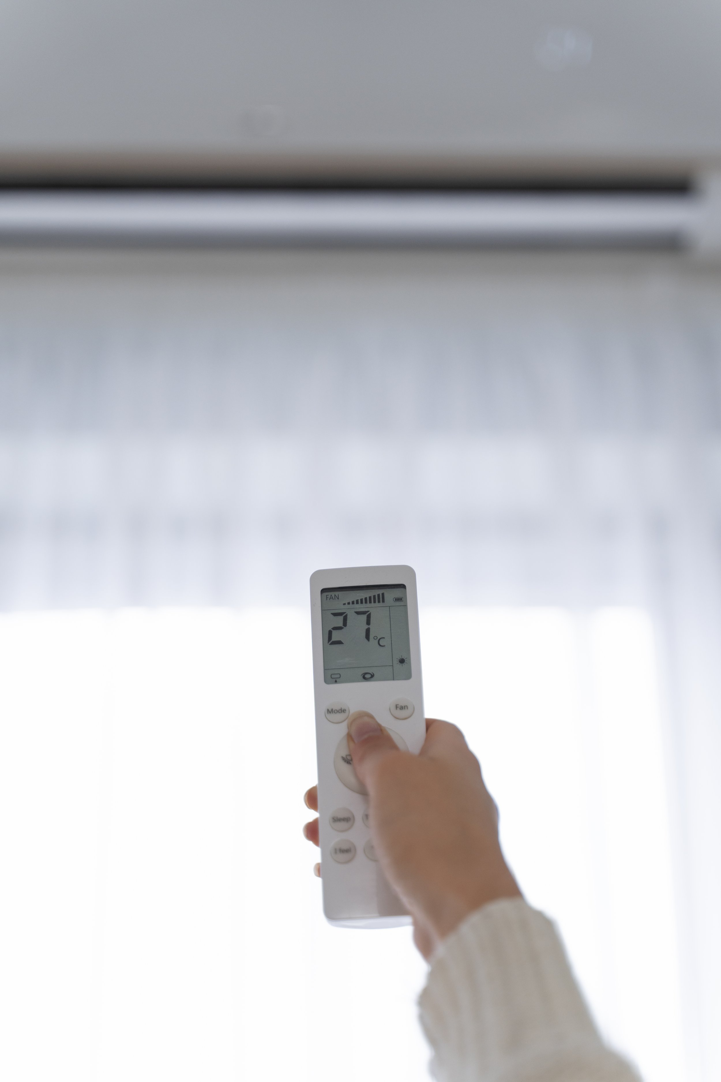 Une main tient une télécommande dirigée vers un aspirateur robot au plafond. L'écran de la télécommande affiche 27°C, probablement une température réglée.
