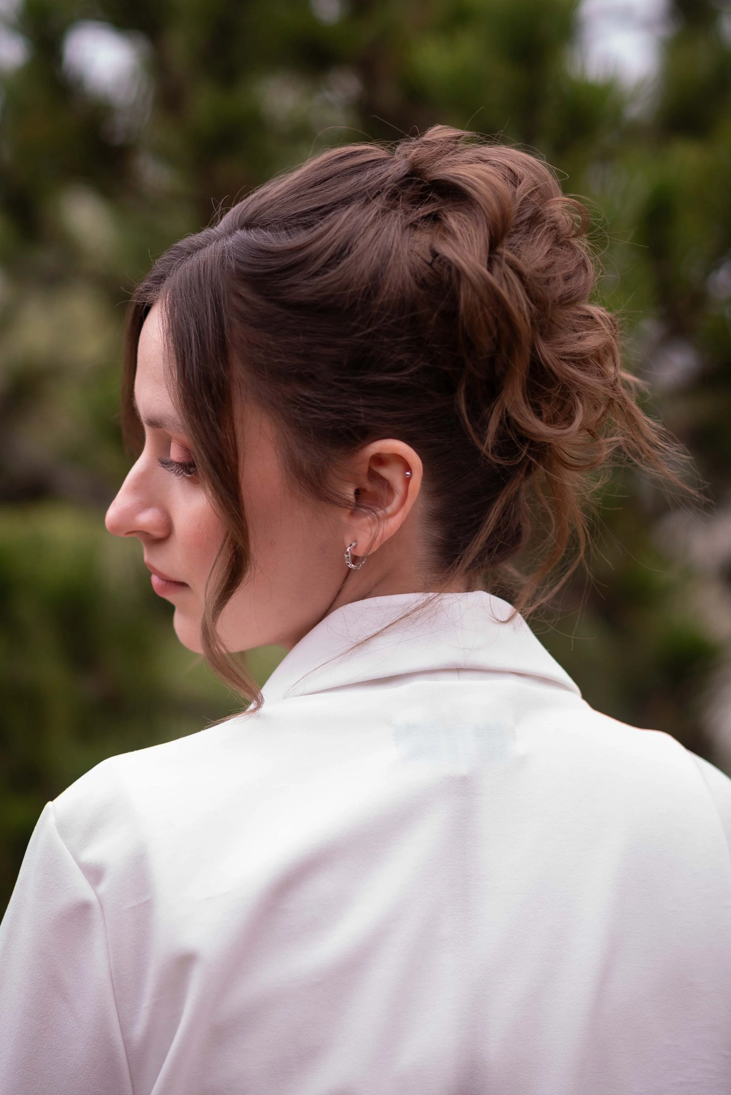 Nahaufnahme einer Braut mit braunen, lockigen Haaren und Ohrpiercing, trägt eine weiße Jacke, im Hintergrund unscharfe grüne Natur.