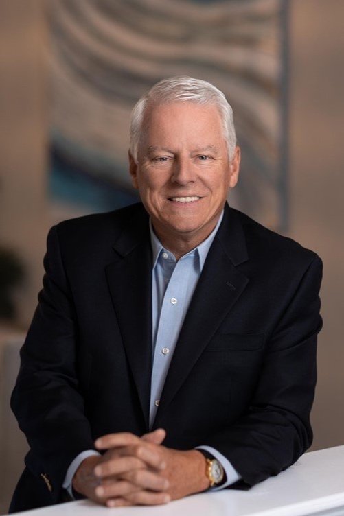 An older man with white hair, wearing a dark suit and light blue shirt, smiling and sitting at a table.