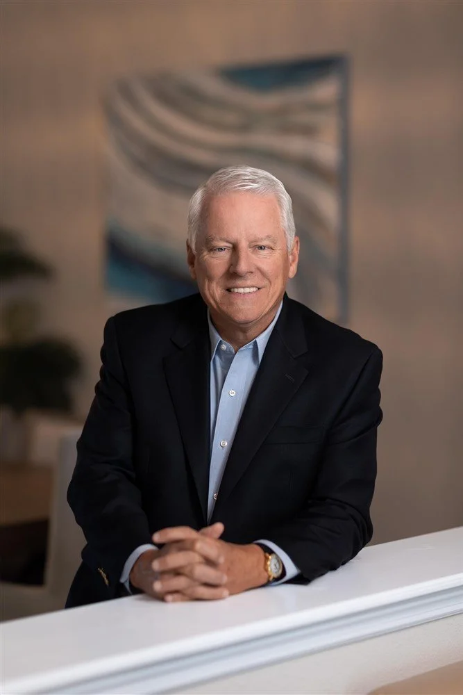A senior man with gray hair in a dark suit and light blue shirt sitting at a white table with hands clasped and smiling, in an office setting with abstract wall art in the background.