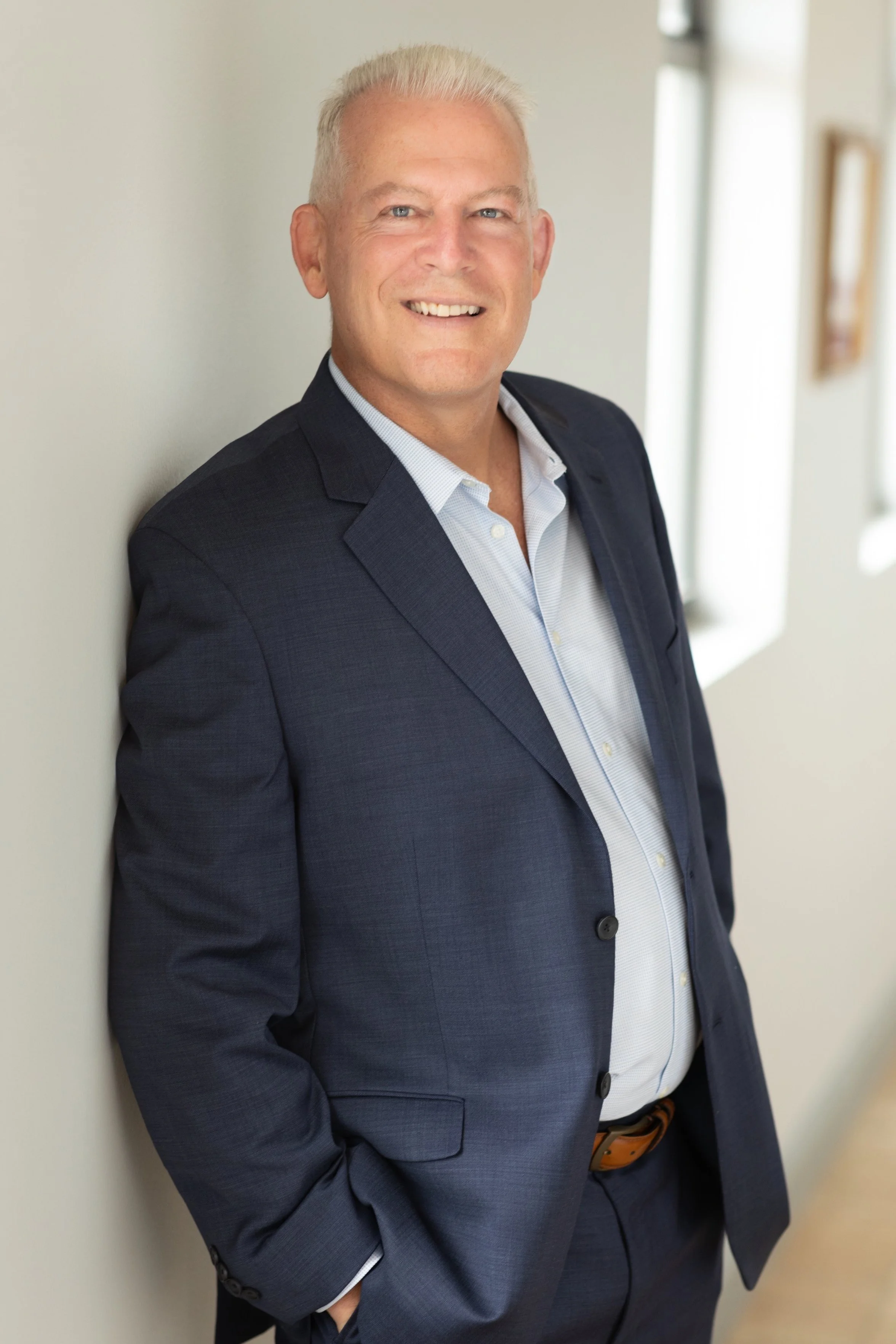 Portrait of a middle-aged man with white hair, dressed in a navy suit and light-colored shirt, smiling and leaning against a wall in a well-lit indoor setting.