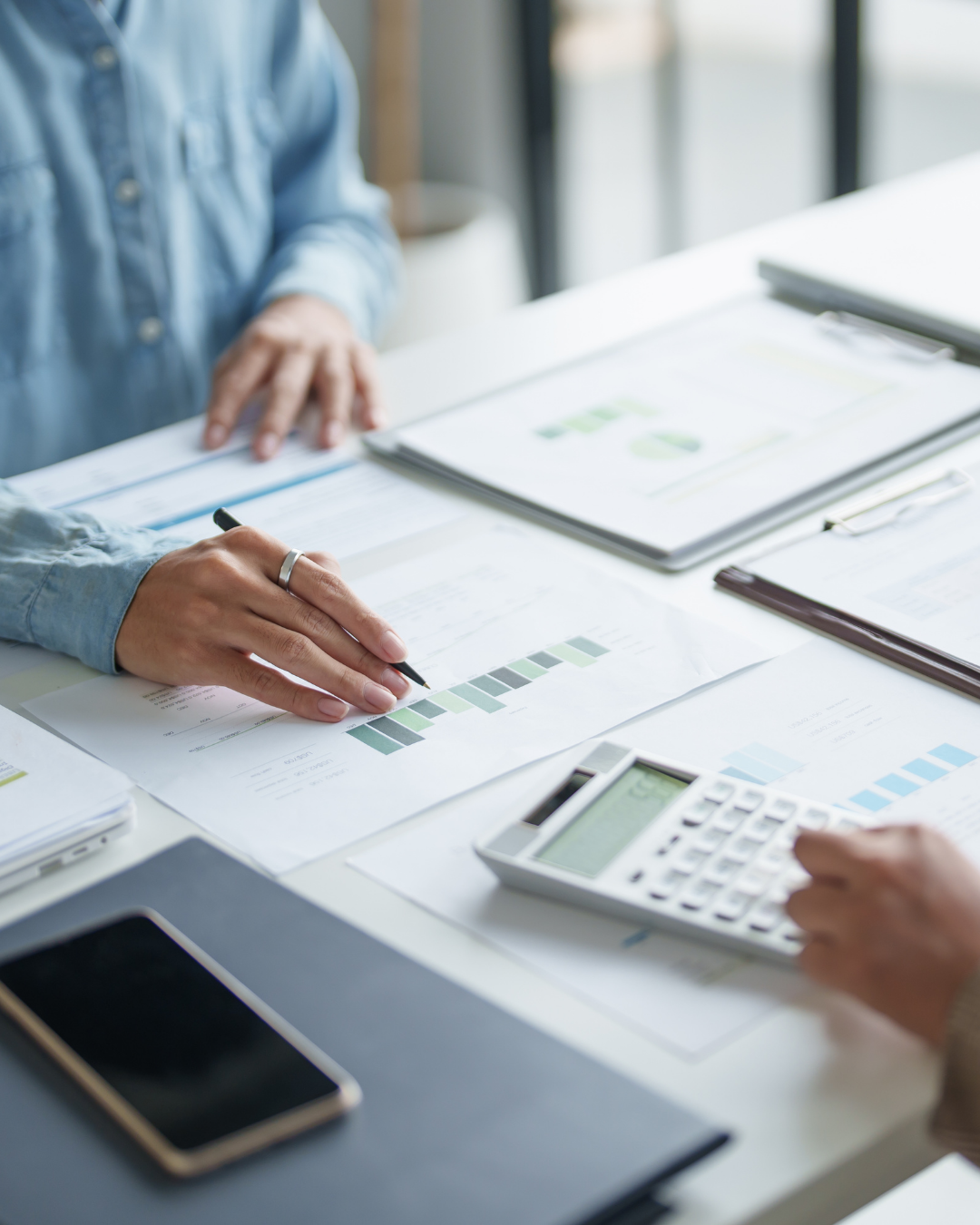Two people working at a desk with documents, charts, a calculator, and a smartphone.