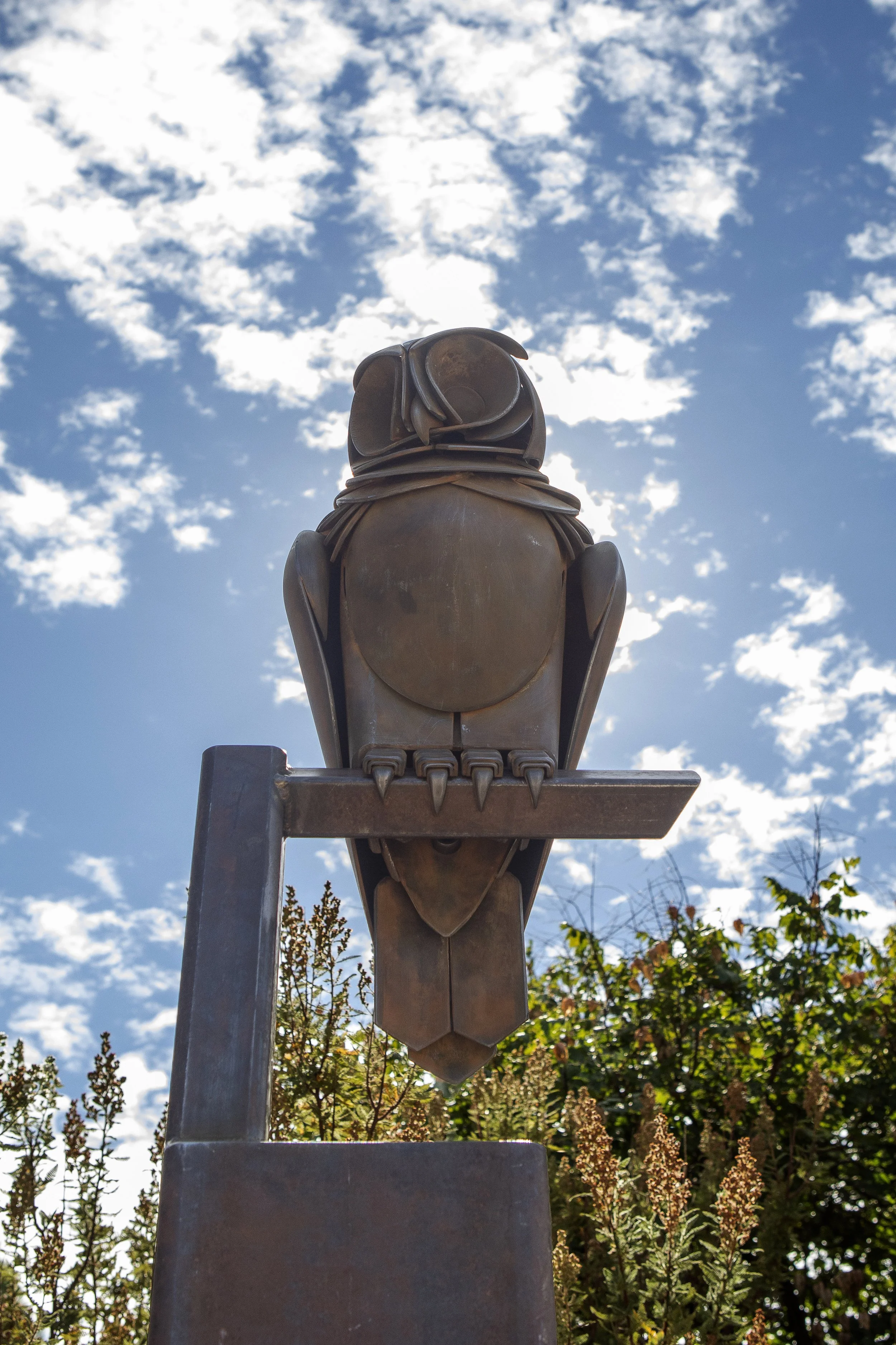 A sculpture of a bird, possibly an owl, made of metal, perched on a stand against a blue sky with scattered clouds, surrounded by trees and plants.