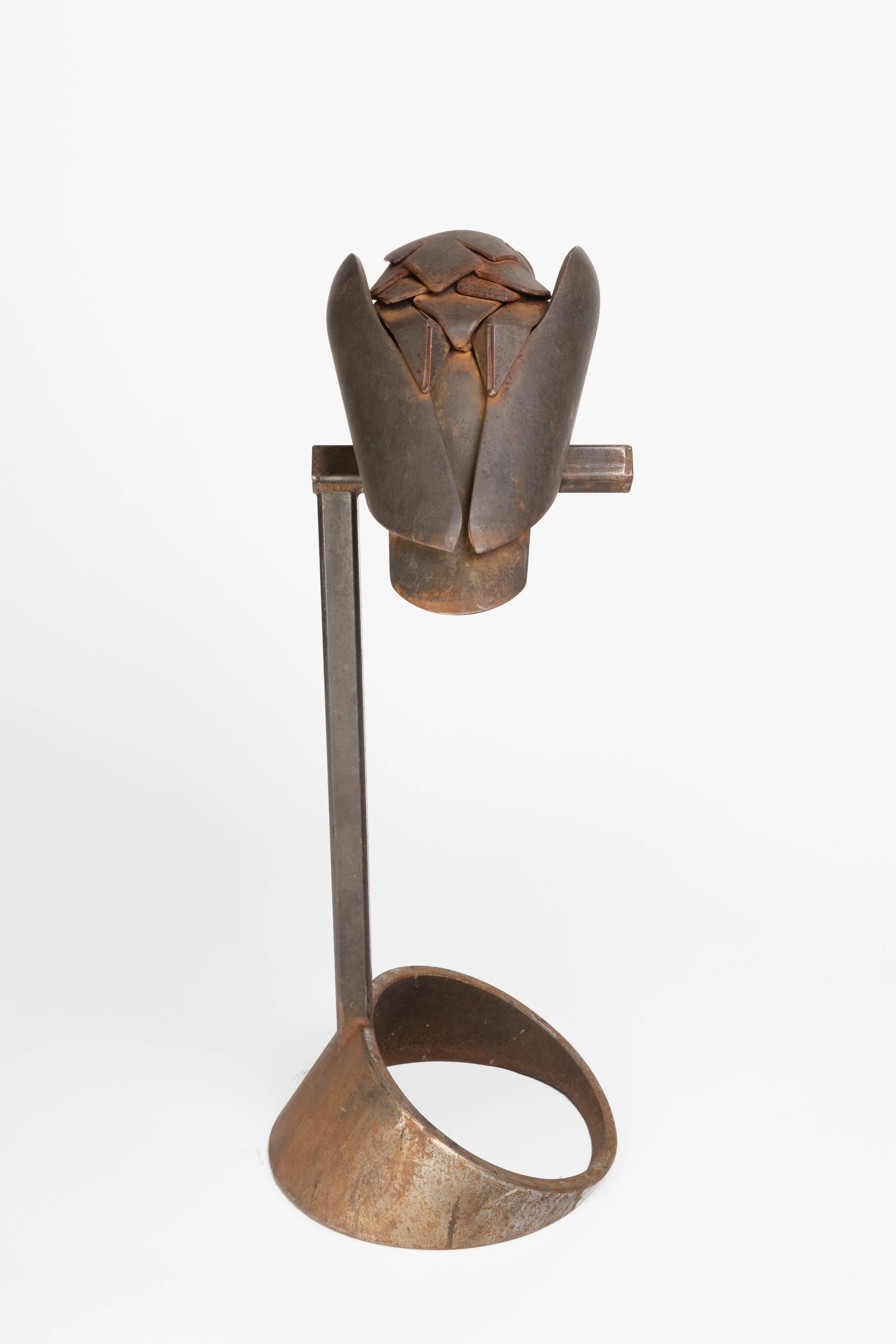 A rustic, metal sculpture of a stylized owl with a domed head made of layered, curved metal plates, mounted on a stand with a curved base.