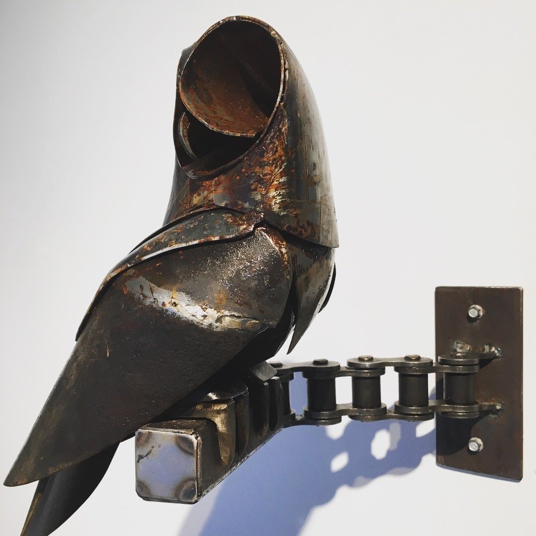 A metal sculpture of an owl mounted on a wall with a chain connecting its body to a rectangular base.