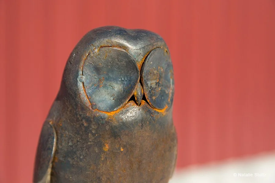 Close-up of a rusty metal owl sculpture with large round eyes against a blurred brick wall background.