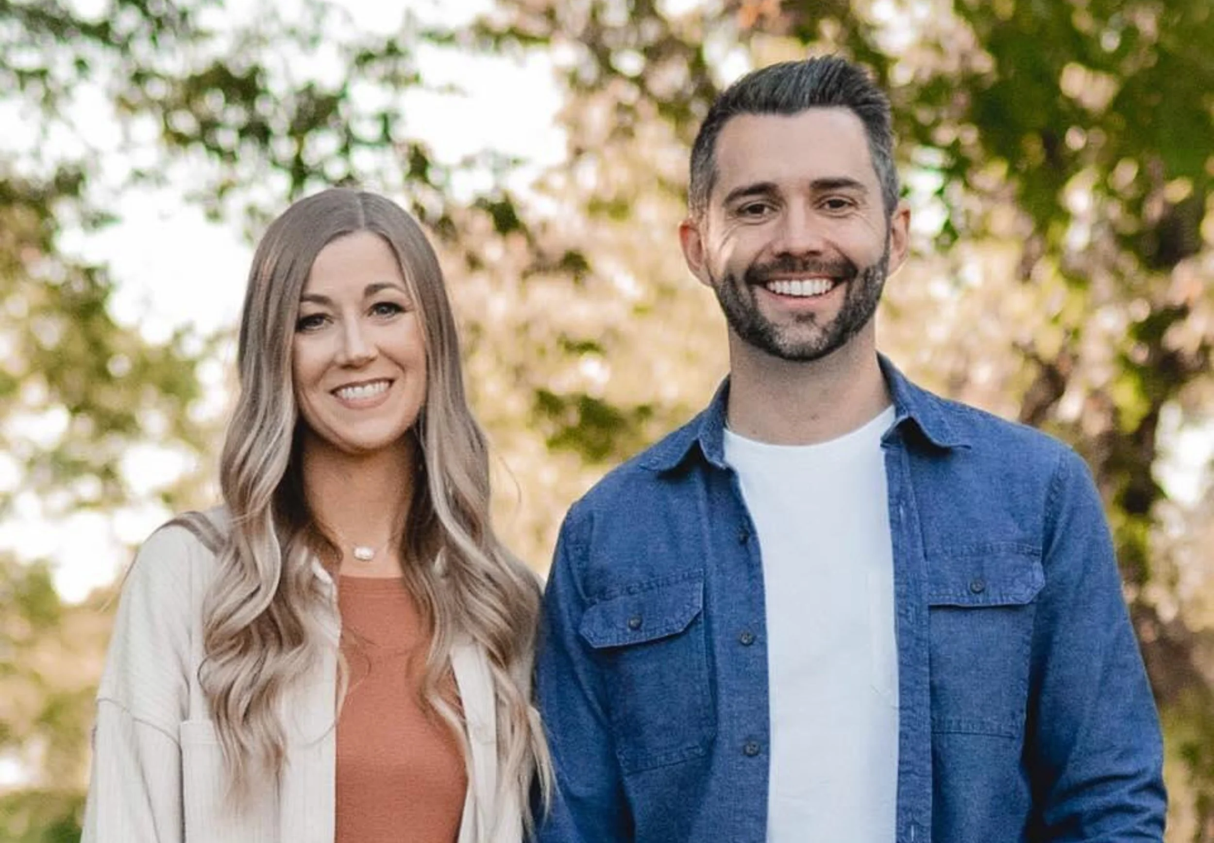 Una pareja sonriente caminando al aire libre en un parque con árboles de fondo.