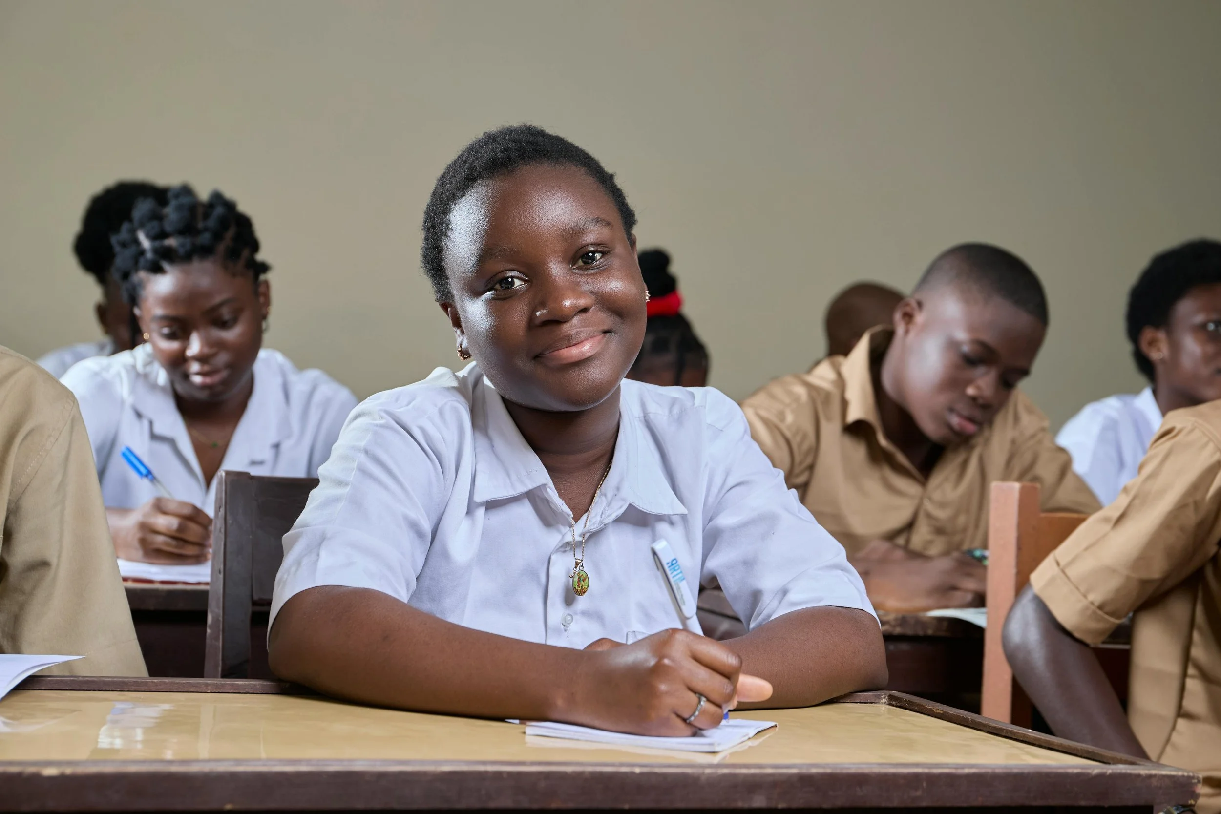 Young Black woman in white shirt smiling and looking at the camera in a classroom, with other students seated around her.