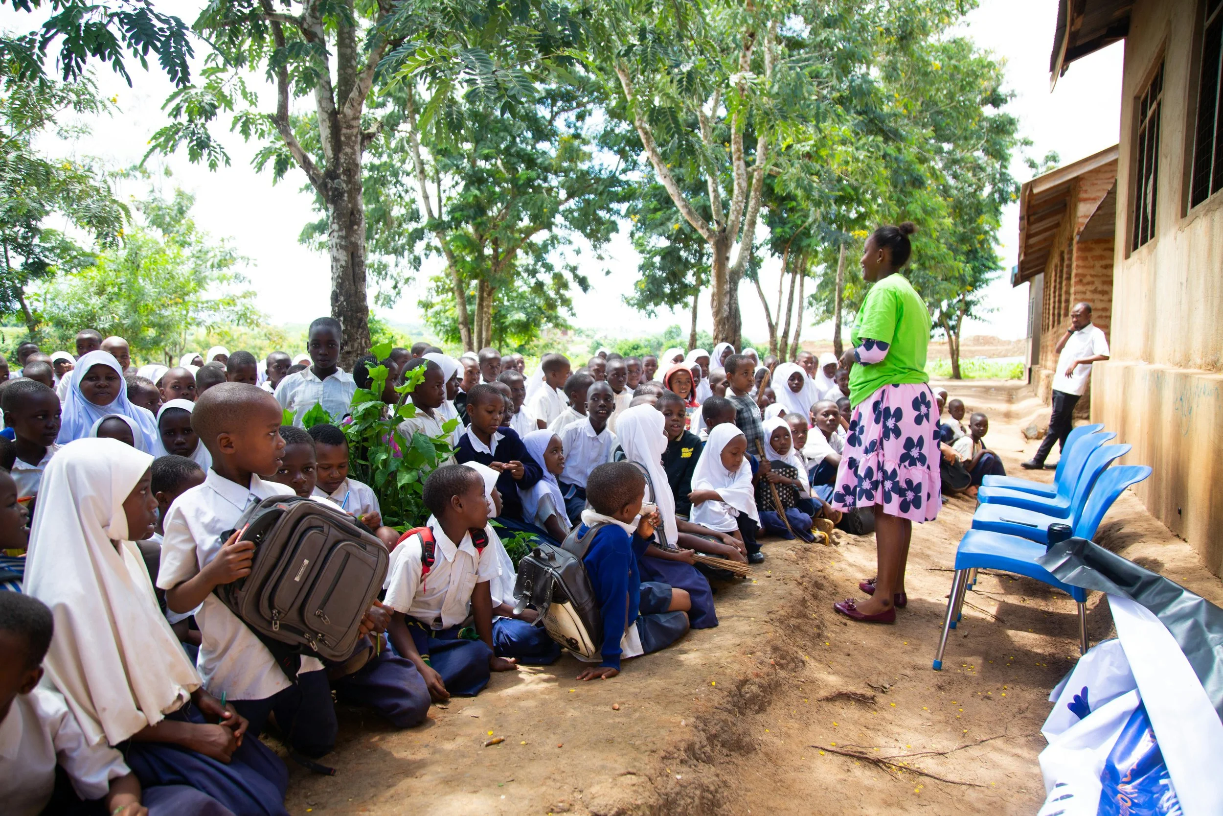A woman in a green shirt and pink skirt addresses a group of children sitting outdoors under trees. The children are of African descent, some wearing school uniforms and head coverings, and appear attentive. There are adults standing in the background near a rustic building.