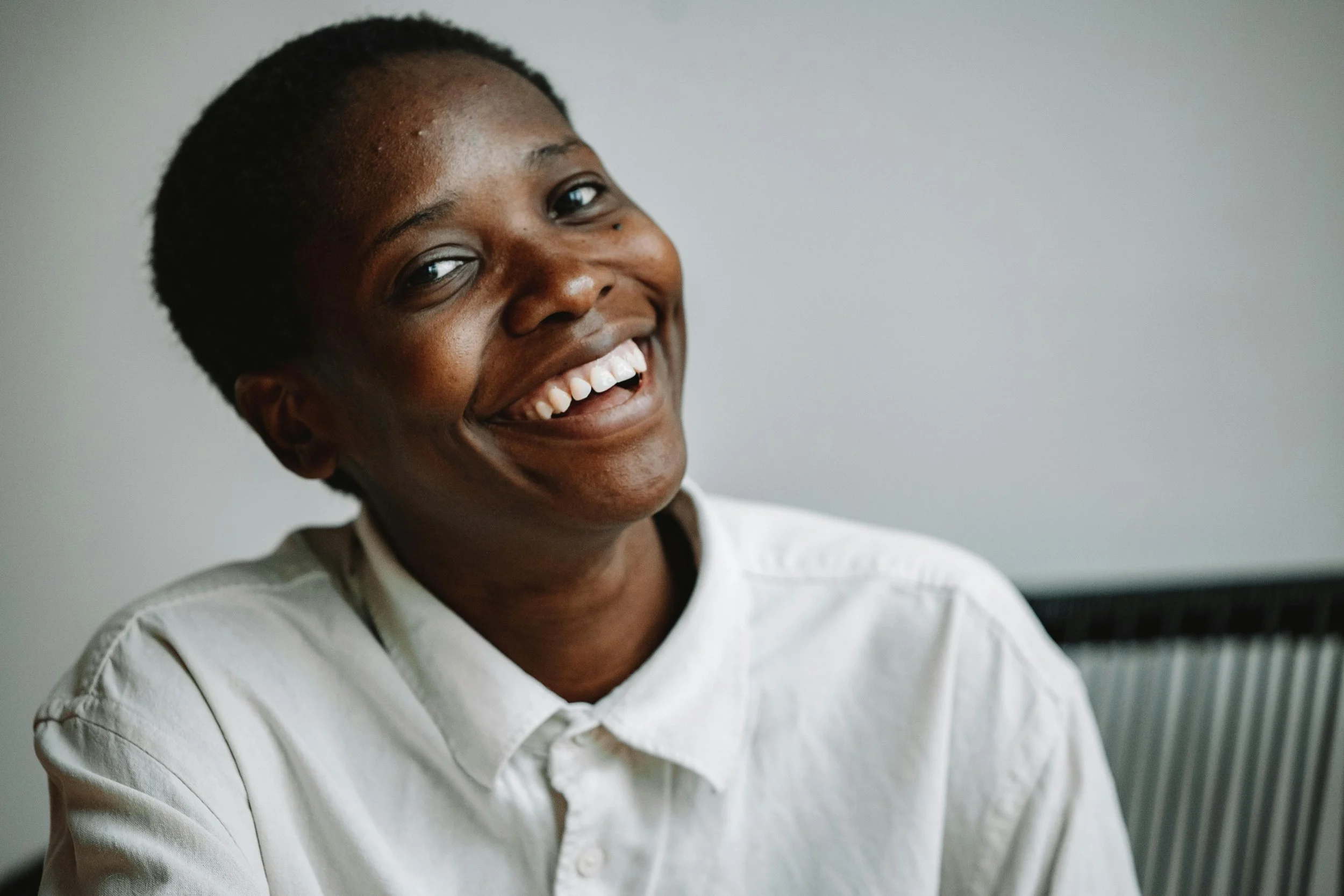 Close-up of a smiling person with short hair, wearing a white shirt, in front of a blurred background.